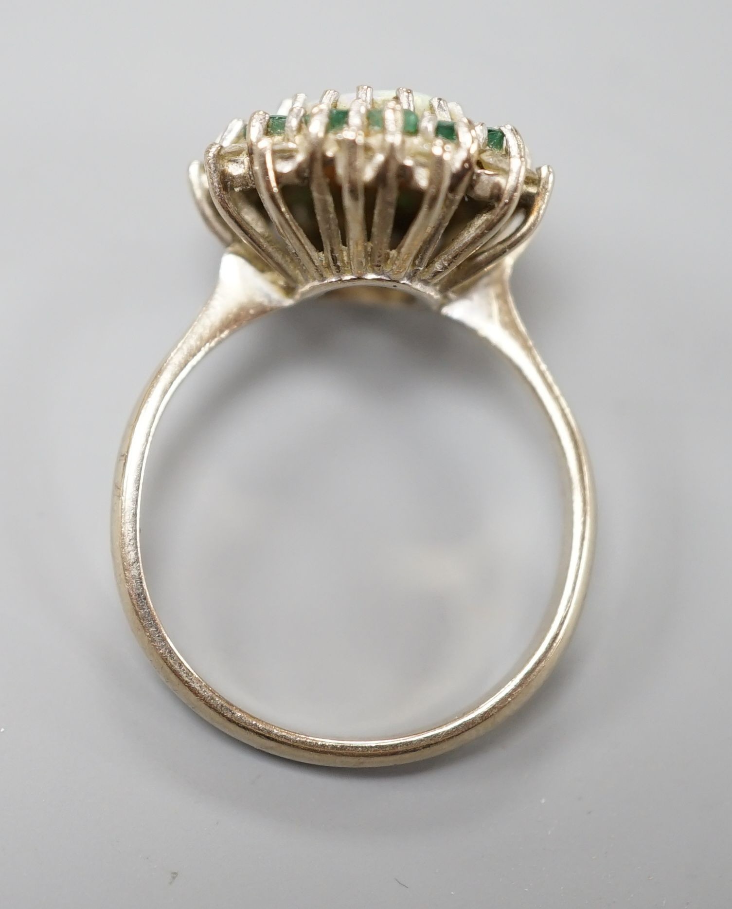 An 18ct white metal, white opal, emerald and diamond set oval cluster ring, size O, gross weight 6.2 grams.